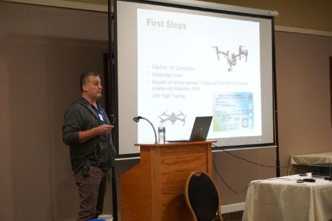 A man beside a projector screen with a picture of a drone and accompanying text.