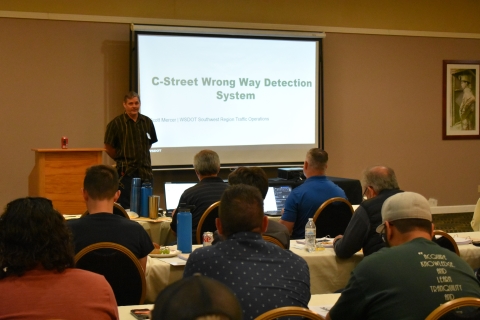 A man stands beside a projector screen in front of an audience. On the screen is the title of the presentation C-Street Wrong Way Detection System.