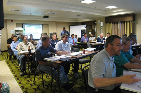 A group of people seated at tables with three ring notebooks in front of them.