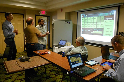 Three men standing and two seated with laptops. The person in the center is describing the software displayed on a screen.
