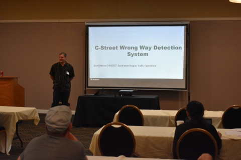 Audience members watch a man presenting beside a projector screen showing a title slide with text C-Street Wrong Way Detection System.