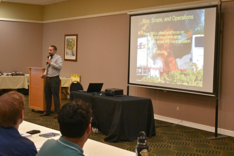A man presents beside a projector screen with text Size, Scope, and Operations. The background of the slide shows a fireball and two vehicles burning.