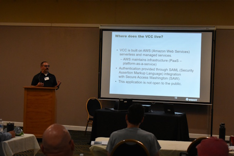 A man gesturing while speaking from a behind a podium. A projector screen displays “Where does the VCC live? VCC is built on AWS …” Slide 19, WSDOT.