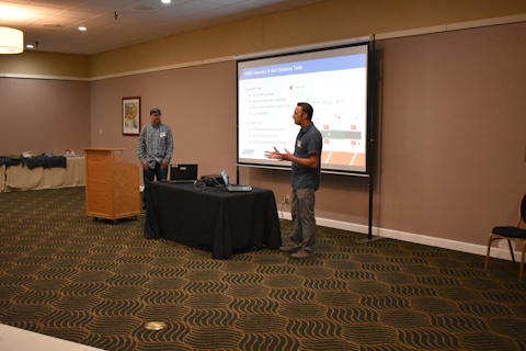 Two men stand on either side of a table with two laptops and a projector, with a projector screen in the background. The man on the left is gesturing while speaking.