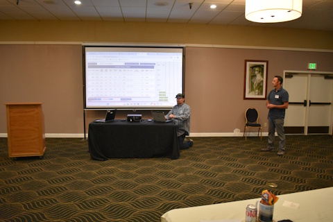A man kneels in front of a laptop on a table. Another man stands to the right side. A projector screen shows a web page with a table of values.