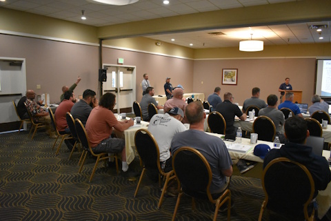 An audience in a classroom style room listens to a speaker in the front. Two others stand to the left. An audience member has his hand in the air to ask a question.