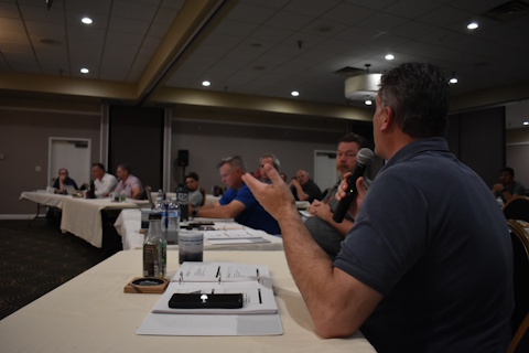 A man seated at a classroom table gestures to the rest of the audience while speaking into a microphone.