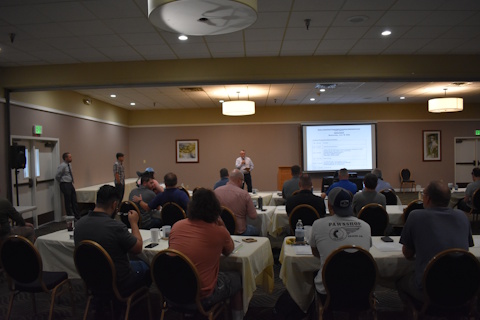 An audience listens to three men speaking in front of a large room next to a projector screen.