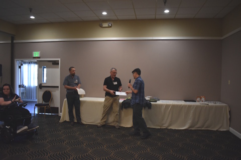 Three men standing in front of a table. The one on the right holds a microphone and is handing a piece of paper to the one in the center. The one on the left holds a piece of paper.