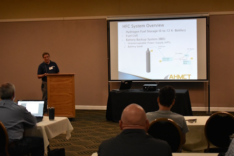 A man speaks standing behind a podium next to a projector screen showing 'HFC System Overview, Hydrogen Fuel Storage, Fuel Cell, Battery Backup System', a picture of a K-bottle, a diagram, and other details.