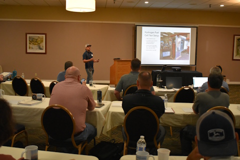 An audience watches a man speaking. A projector screen displays “Hydrogen Fuel Cell Test Setup” and an image of an HFC in a cabinet in a lab.