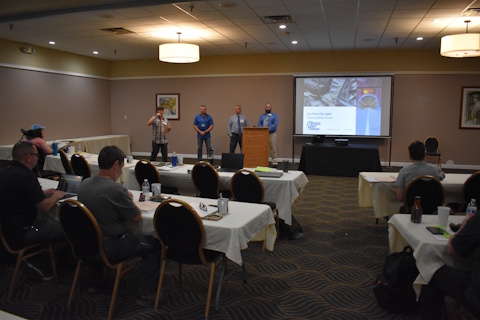 Four men stand beside a projector screen in front of an audience seated at tables. The one on the left is speaking into a microphone. On the screen is a title slide for the presentation.