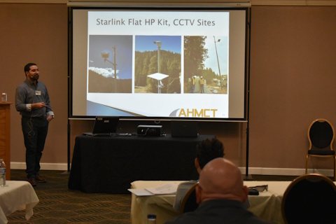 A man stands to the left of a projector screen displaying “Starlink Flat HP Kit, CCTV Sites” and three photos of CCTV camera sites with the Starlink panels mounted on the same poles.