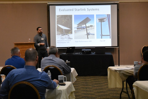 A man stands between a podium and a projector screen with Evaluated Starlink Systems and three pictures showing panels mounted on poles.