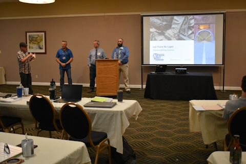 Four men stand beside a projector screen with a presentation title slide. The person on the left is speaking about the others. Tables in front.