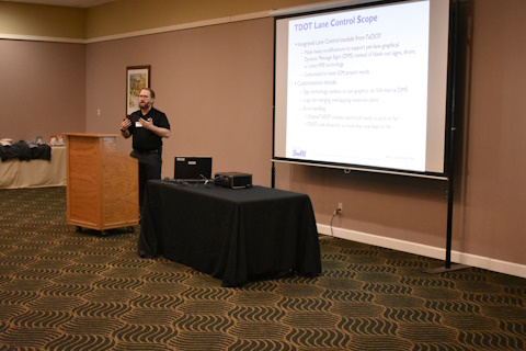 A man gestures while speaking next to a podium and projector screen.