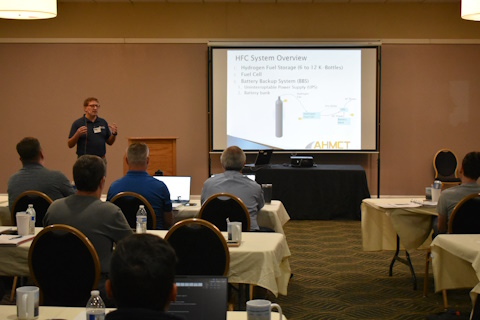 A man gestures while presenting beside a podium and projector screen with a slide showing HFC System Overview and a cylinder.