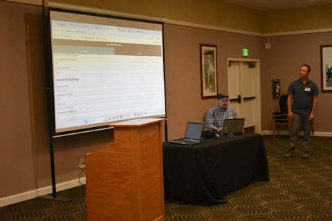 One man seated at a table in front of a laptop beside a projector screen. Another man standing to the right looking at the screen.