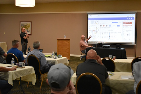 A man seated in front of a laptop and a projector screen gestures to the screen while speaking to an audience. Another man stands to the side.