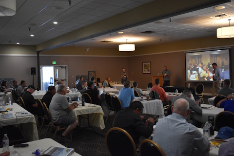 An audience listens to three men standing in the front of room with a projector screen and podium.